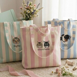May include: Three striped tote bags with embroidered pet portraits. One bag is light blue and white with a cat and the name "Muffins". Another is pink and white with two cats and the names "Grace & Frankie". The third is blue and white with a dog and the name "Cobalt".