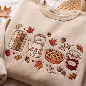 May include: A beige crewneck sweatshirt featuring embroidered autumn-themed motifs including a stack of three books tied with string, a floral teapot, a lattice-crust pie, and a jar labelled JAM, surrounded by scattered acorns, berries, and leaves.