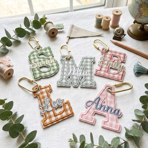 May include: Five fabric letter keychains featuring a gingham pattern in green, gray, pink, orange, and light pink. Each letter is embroidered with a name: Sarah, Mia, Emily, Leo, and Anna. The keychains include gold-toned metal lobster clasps.