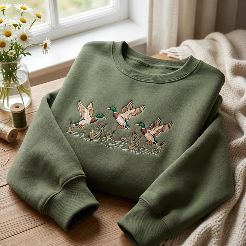 Może przedstawiać: Zielona bluza w kolorze oliwkowym z haftowanym wzorem trzech kaczek w locie nad trzcinami i wodą. Bluza ma okrągły dekolt i prążkowane mankiety. Wz&oacute;r jest szczeg&oacute;łowy z brązowymi, zielonymi i beżowymi kolorami.