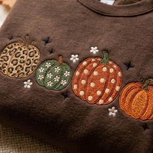 Op de afbeelding: Een donkerbruin sweatshirt met geborduurde pompoenontwerpen op de borst. De pompoenen bevatten een luipaardprint, een groene pompoen met witte bloemen, een oranje pompoen met witte stippen en een effen oranje pompoen.
