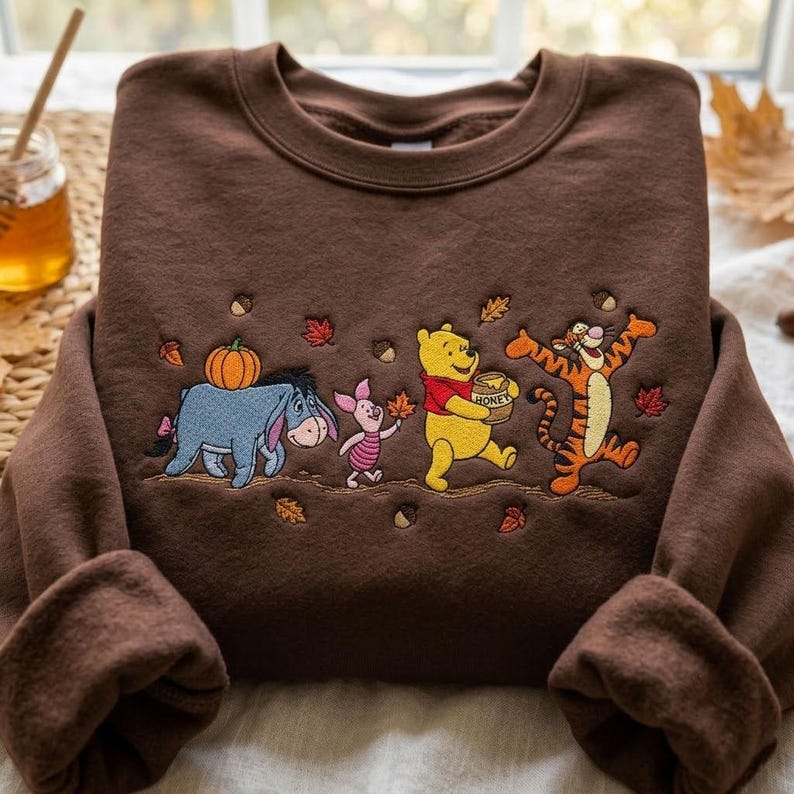 May include: A dark brown crewneck sweatshirt featuring an embroidered design of Eeyore carrying a pumpkin, Piglet holding a leaf, Winnie the Pooh with a honey pot, and Tigger, all surrounded by scattered autumn leaves and small acorns.