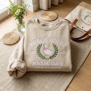 May include: Beige sweatshirt with embroidered "PORCH GOOSE SOCIAL CLUB" design. Features a goose wearing a pink bow within a laurel wreath. Includes the text "EST. 1983" and "Gotta go dress my goose". A matching tote bag is visible.