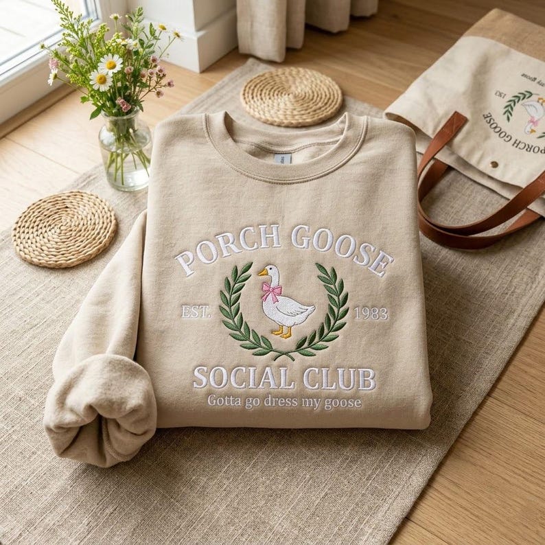 May include: Beige sweatshirt with embroidered "PORCH GOOSE SOCIAL CLUB" design. Features a goose wearing a pink bow within a laurel wreath. Includes the text "EST. 1983" and "Gotta go dress my goose". A matching tote bag is visible.