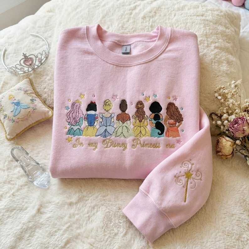 May include: Pink sweatshirt with embroidered princess design and the text "In my Disney Princess era." The sleeve features a gold embroidered wand. A tiara, glass slipper, and embroidered pillow are also visible.
