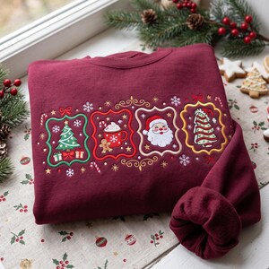 K&ouml;nnte beinhalten: Ein kastanienbraunes Sweatshirt mit vier gestickten rechteckigen Rahmen mit gewelltem Rand. Die Motive zeigen einen Weihnachtsbaum mit Geschenken, eine Tasse hei&szlig;e Schokolade mit einem Lebkuchenmann, ein Weihnachtsmann-Gesicht und einen geschm&uuml;ckten Baum.