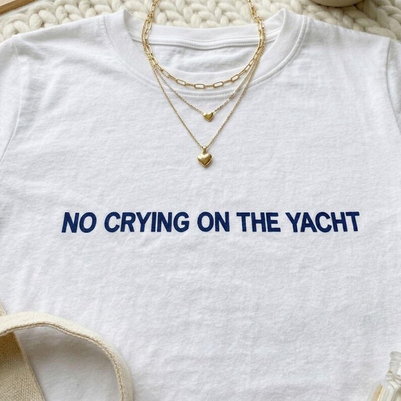Puede incluir: Camiseta blanca con el texto azul marino "NO CRYING ON THE YACHT". Un collar dorado en capas con colgantes de coraz&oacute;n est&aacute; sobre la camiseta. La camiseta est&aacute; sobre una superficie blanca.