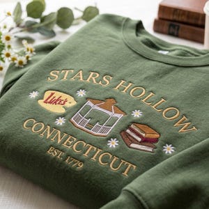 May include: An olive green crewneck sweatshirt featuring embroidered text that reads Stars Hollow Connecticut Est. 1779. The design includes a yellow Luke's sign, a white gazebo, a stack of three books, and small white daisy flowers.