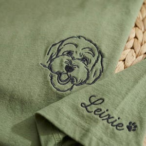 May include: Olive green fabric with an embroidered dog face and the name "Leixie" in cursive. A small paw print is next to the name. The dog's face is detailed with dark blue thread.