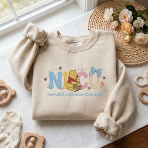 May include: A beige sweatshirt with the embroidered word "NICU" in pastel colors, featuring a cartoon bear and bees. The text "neonatal intensive care unit" is below the embroidered design. The sweatshirt has long sleeves.