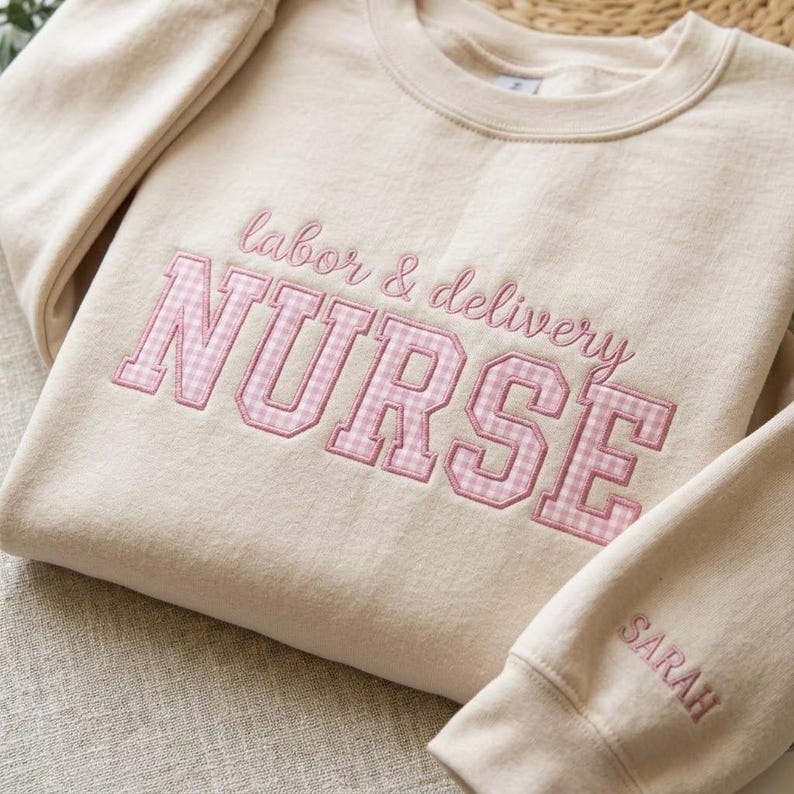 May include: A beige sweatshirt with the words "labor & delivery NURSE" embroidered in pink. The word "NURSE" is in a pink gingham pattern. The sleeve cuff has the name "SARAH" embroidered in pink.