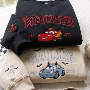 May include: A black sweatshirt featuring an embroidered red race car with a witch hat, the text MCQUEEN EST. 1977, and spiderwebs. A beige sweatshirt featuring an embroidered blue car with horns, the text SALLY EST. 2002, and spiderwebs.