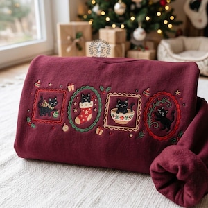 May include: A deep burgundy sweatshirt featuring four embroidered rectangular and oval frames containing black cats in festive holiday scenes, including a cat with a scarf, a cat in a stocking, a cat in a bowl, and a cat with holly branches.