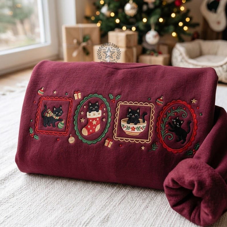 May include: A deep burgundy sweatshirt featuring four embroidered rectangular and oval frames containing black cats in festive holiday scenes, including a cat with a scarf, a cat in a stocking, a cat in a bowl, and a cat with holly branches.