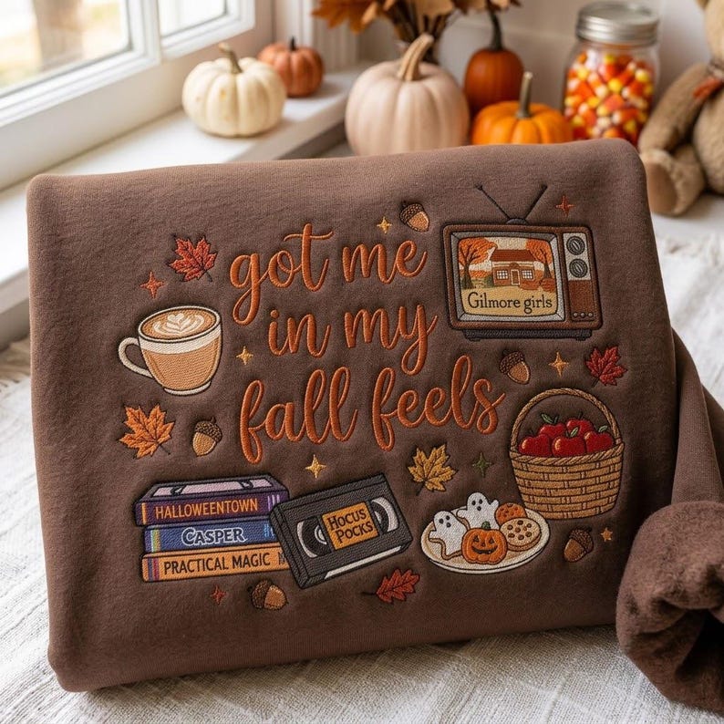 May include: A brown sweatshirt featuring embroidered autumn-themed designs and the text "got me in my fall feels". The embroidery includes a coffee mug, a television displaying "Gilmore girls", a stack of VHS tapes, a basket of apples, and cookies.
