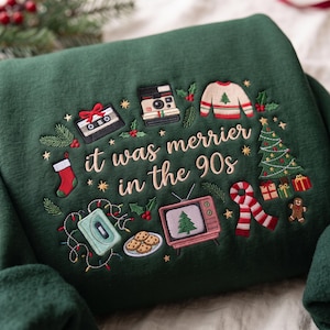 Peut inclure: Un sweat-shirt vert foncé avec des broderies sur le thème de Noël et des années 90, incluant une cassette, un appareil photo instantané, une télévision et des biscuits. Le texte "it was merrier in the 90s" est entouré d'un sapin et d'une écharpe.