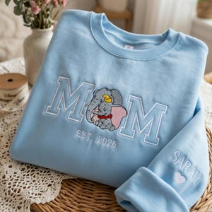 Puede incluir: Sudadera azul claro con la palabra "MOM" bordada en blanco, con un gráfico de elefante. El puño de la manga tiene el nombre "SARAH" y un corazón. El texto "EST. 2026" está bordado debajo del gráfico del elefante.