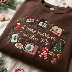 May include: A dark brown crewneck sweatshirt featuring embroidered Christmas-themed designs and the text "it was merrier in the 90s". The embroidery includes a cassette tape, a camera, a stocking, a sweater, a television, a scarf, and a tree.