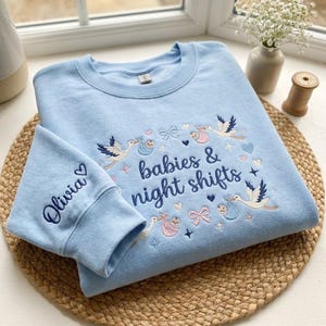 May include: Light blue sweatshirt with embroidered design. The design includes storks carrying babies, bows, hearts, and the text "babies & night shifts." The left sleeve has the name "Olivia" embroidered with a heart.