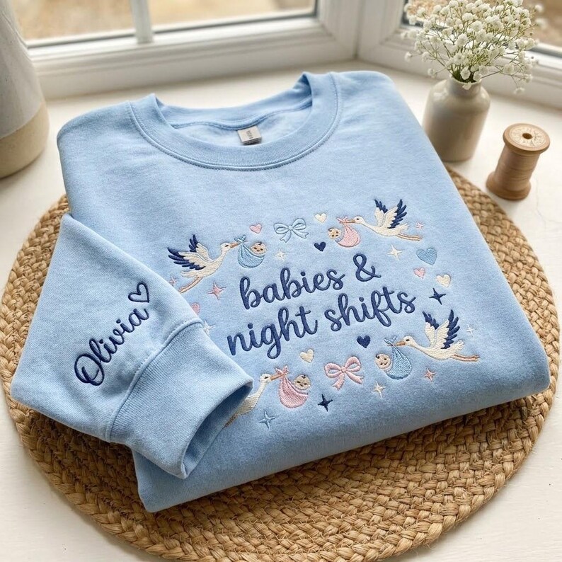 May include: Light blue sweatshirt with embroidered design. The design includes storks carrying babies, bows, hearts, and the text "babies & night shifts." The left sleeve has the name "Olivia" embroidered with a heart.