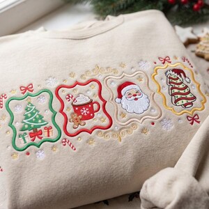 K&ouml;nnte beinhalten: Ein beigefarbenes Sweatshirt mit vier gestickten rechteckigen Rahmen mit gewellten R&auml;ndern. Die Motive zeigen einen gr&uuml;nen Weihnachtsbaum, eine rote Tasse mit hei&szlig;er Schokolade und Lebkuchenmann, ein Weihnachtsmann-Gesicht und einen Baum.