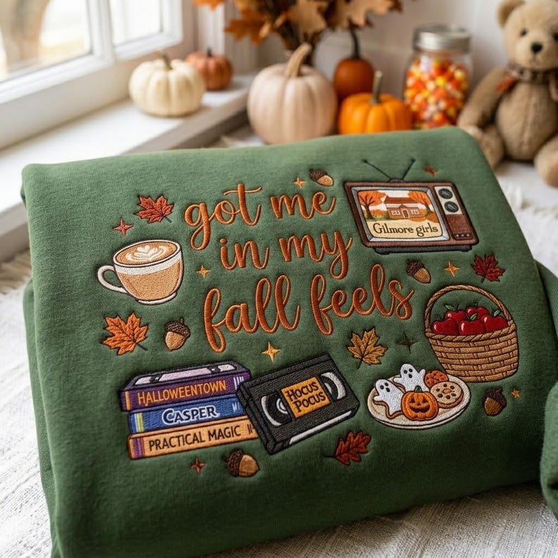 May include: An olive green sweatshirt featuring embroidered autumn-themed designs and the text "got me in my fall feels". The embroidery includes a coffee mug, a television showing "Gilmore girls", a basket of apples, VHS tapes, and cookies.