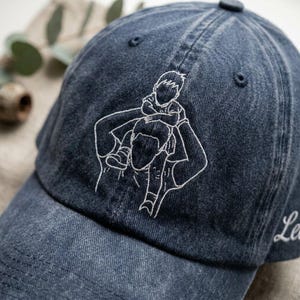 May include: A denim blue baseball cap with a white embroidered design of a person carrying a child on their shoulders. The cap has a curved brim and a small, centered button on top. The word "Le" is embroidered on the side.