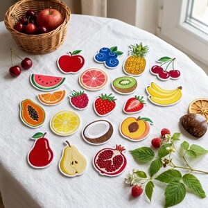 Op de afbeelding: Een verzameling geborduurde fruitpatches met een rode appel, bosbessen, ananas, kersen, watermeloenplak, grapefruitplak, kiwi, bananen, framboos, aardbei, papaja, sinaasappelplak, citroenplak, kokosnoot, perzik, peer en een granaatappel.