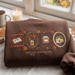 Puede incluir: Una sudadera marrón oscuro con bordados de temática otoñal que incluyen un libro abierto con gafas, un jarrón con hojas, un tarro y una pequeña casa, todo en marcos decorativos con el texto "cozy season" bordado en la parte inferior.