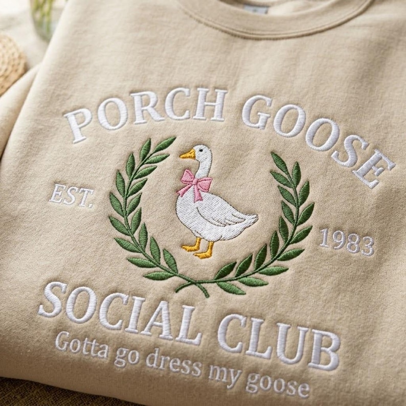 May include: Beige sweatshirt with embroidered design. Features a white goose wearing a pink bow, surrounded by a green wreath. Text includes "PORCH GOOSE SOCIAL CLUB" and "Gotta go dress my goose".