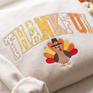 Op de afbeelding: Een crèmekleurig sweatshirt met het woord THANKFUL geborduurd in een college-lettertype met diverse gele en oranje patronen. Onder de tekst staat een geborduurde kalkoen met een zwarte hoed en kleurrijke veren in de staart.