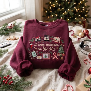 May include: A maroon crewneck sweatshirt featuring embroidered 90s-themed Christmas graphics and the text "it was merrier in the 90s". The design includes a cassette tape, instant camera, television, Christmas tree, and cookies on a light fabric.