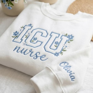 May include: White sweatshirt with blue embroidered text and floral accents. The text reads "ICU nurse" with a floral design. The sleeve cuff features the name "Olivia" in blue script. The sweatshirt is made of a soft material.