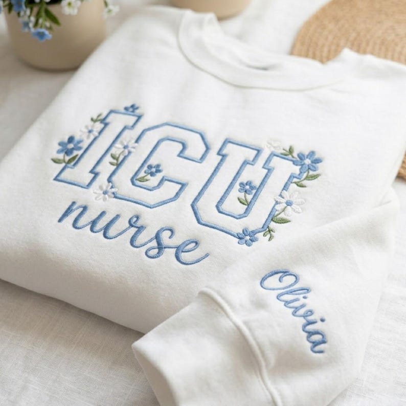 May include: White sweatshirt with blue embroidered text and floral accents. The text reads "ICU nurse" with a floral design. The sleeve cuff features the name "Olivia" in blue script. The sweatshirt is made of a soft material.