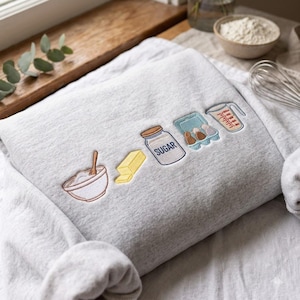 Könnte beinhalten: Ein hellgrau meliertes Sweatshirt mit einer Stickerei von Backzutaten, darunter eine Rührschüssel mit Löffel, ein Stück Butter, ein mit SUGAR beschriftetes Glas, ein Eierkarton und ein Messbecher, die in einer horizontalen Reihe angeordnet sind.