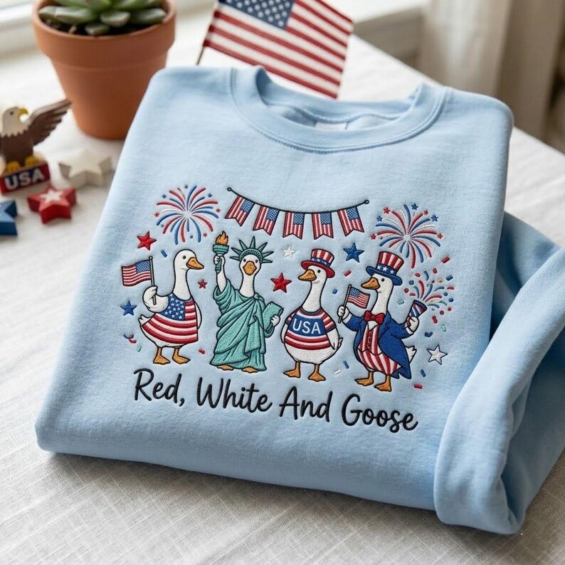 May include: Light blue sweatshirt with an embroidered design featuring geese dressed in patriotic attire, including a Statue of Liberty goose. The text "Red, White And Goose" is below the design. Fireworks and American flags complete the design.