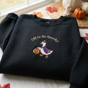 May include: A black crewneck sweatshirt featuring an embroidered design of a white duck wearing a purple cape and a black witch hat. The duck holds an orange jack-o'-lantern bucket. The text Off to Be Spooky is embroidered in an arc above.