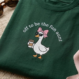 May include: Dark green t-shirt with an embroidered design of a white goose wearing sunglasses and a pink bow, holding a coffee cup. The text "off to be the fun aunt" is arched above the goose.