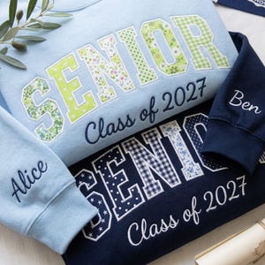 Peut inclure: Un sweat-shirt bleu clair avec le mot SENIOR en appliqué floral et vichy vert, et un sweat-shirt bleu marine avec le mot SENIOR en appliqué étoilé et vichy bleu. Les deux portent le texte Class of 2027 et des prénoms brodés sur les manches.