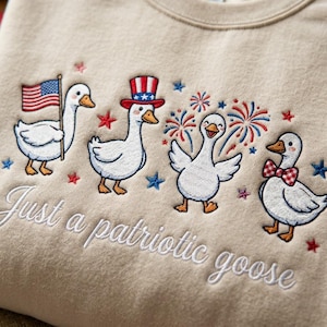 May include: Beige sweatshirt with embroidered patriotic geese. One goose holds an American flag, another wears a top hat, and a third has a bow tie. The text "Just a patriotic goose" is embroidered below.