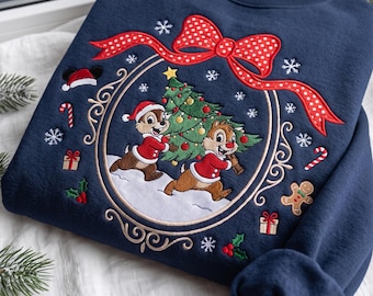 May include: A navy blue crewneck sweatshirt featuring an embroidered design of two chipmunks carrying a decorated Christmas tree. The scene is framed by a gold oval border topped with a large red polka-dot bow, surrounded by snowflakes, candy canes, gifts, and a gingerbread man.