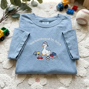 K&ouml;nnte beinhalten: Hellblaues T-Shirt mit dem Schriftzug "outnumbered by boys" und einer Cartoon-Ente mit Baseballkappe. Das Shirt zeigt gestickte Bilder von einem Lastwagen, einem Auto, einem Fu&szlig;ball, einem Schraubenschl&uuml;ssel und Bausteinen.