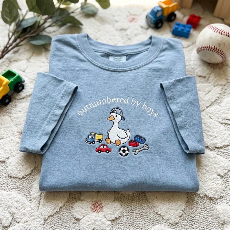 K&ouml;nnte beinhalten: Hellblaues T-Shirt mit dem Schriftzug "outnumbered by boys" und einer Cartoon-Ente mit Baseballkappe. Das Shirt zeigt gestickte Bilder von einem Lastwagen, einem Auto, einem Fu&szlig;ball, einem Schraubenschl&uuml;ssel und Bausteinen.