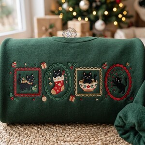 May include: A dark green sweatshirt featuring four embroidered frames with black cats. The designs include a cat with a scarf and ornament, a cat in a Christmas stocking, a cat in a bowl, and a cat amidst greenery. Small gifts and ornaments are scattered between the frames.