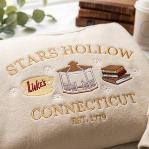 May include: A beige sweatshirt featuring gold embroidered text that reads Stars Hollow Connecticut Est. 1779. The design includes a yellow coffee cup with the red text Luke's, a white gazebo, a stack of three books, and small white daisy flowers.