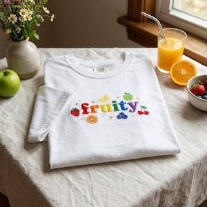 May include: A white t-shirt featuring the word "fruity" embroidered in colorful, puffy letters across the chest. The design is surrounded by small embroidered fruit icons, including a strawberry, orange slice, apple, blueberries, and cherries.