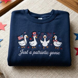 May include: Navy blue sweatshirt featuring an embroidered design of four patriotic geese. The geese are adorned with an American flag, a top hat, fireworks, and a bow tie. The text "Just a patriotic goose" is embroidered below the geese.
