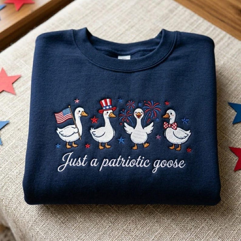 May include: Navy blue sweatshirt featuring an embroidered design of four patriotic geese. The geese are adorned with an American flag, a top hat, fireworks, and a bow tie. The text "Just a patriotic goose" is embroidered below the geese.