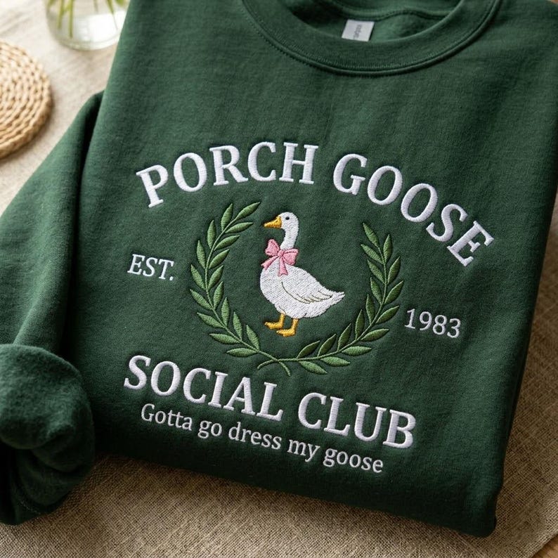 May include: A dark green sweatshirt featuring white embroidered text and a goose design. The text says "PORCH GOOSE SOCIAL CLUB", "EST. 1983", and "Gotta go dress my goose". The goose wears a pink bow.