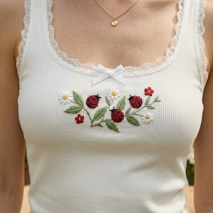 Könnte beinhalten: Ein cremefarbenes, geripptes Tanktop mit zarter Spitzenborte am Rundhalsausschnitt und einer kleinen weißen Schleife in der Mitte. Der Brustbereich ist mit einer Stickerei aus roten Marienkäfern, grünen Blättern und weißen Blumen verziert.